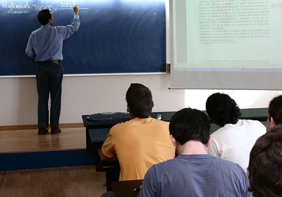Un profesor escribiendo en la pizarra mientras da clase y estudiantes tomando apuntes