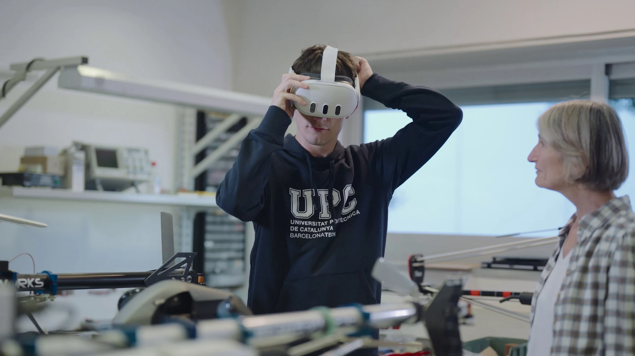 En un laboratori, un estudiant de la UPC s’ajusta un visor de realitat virtual mentre l’observa una professora.